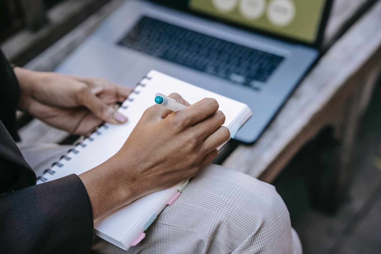 Hands writing in a notebook beside a laptop