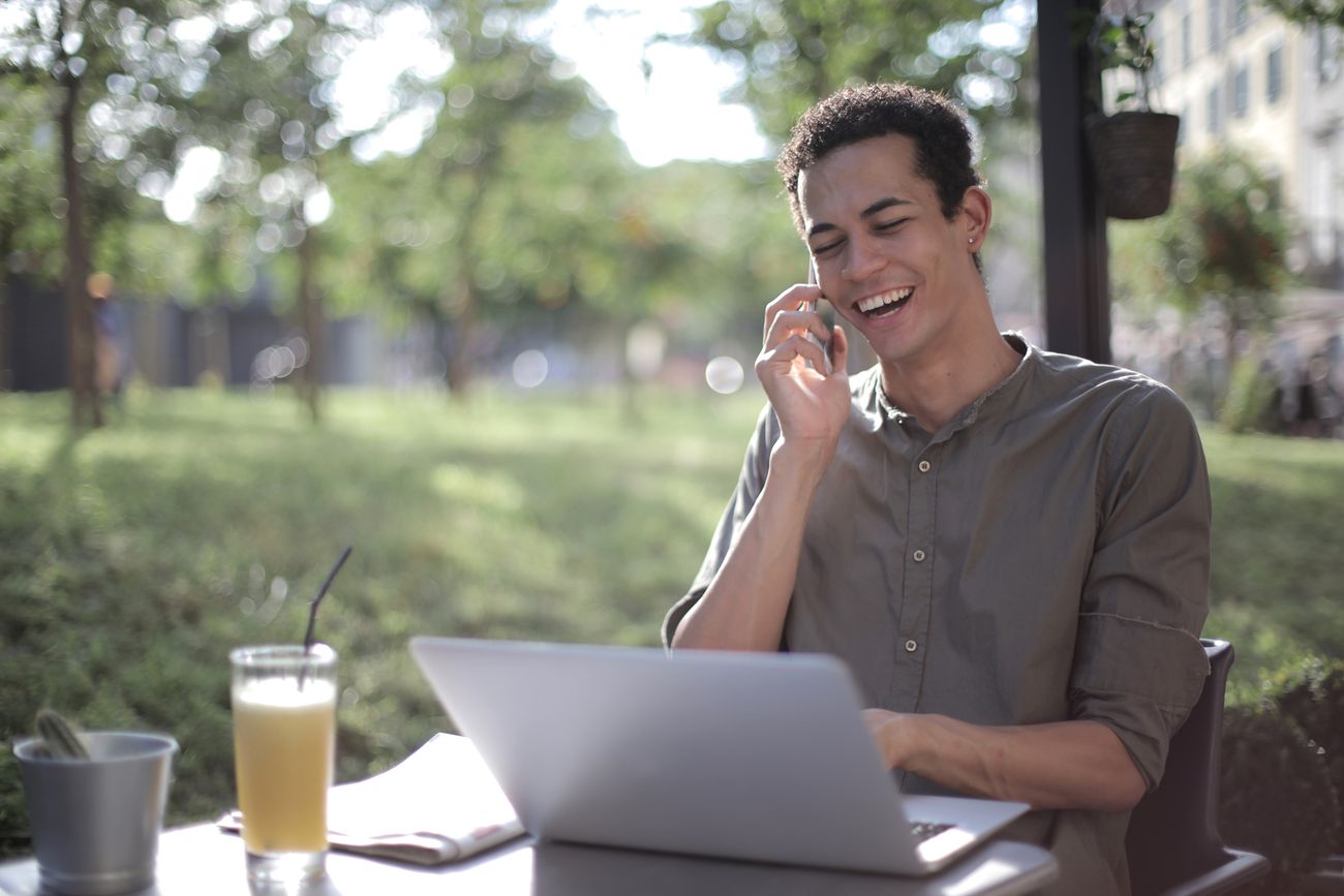 A man laughing on the phone while working on a laptop outdoors